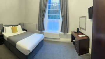 A hotel room with a bed, bedside tables, a desk, a chair, a window with curtains, a radiator, and a TV.