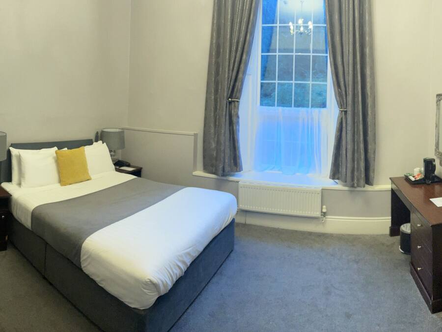 A hotel room with a bed, bedside tables, a desk, a chair, a window with curtains, a radiator, and a TV.