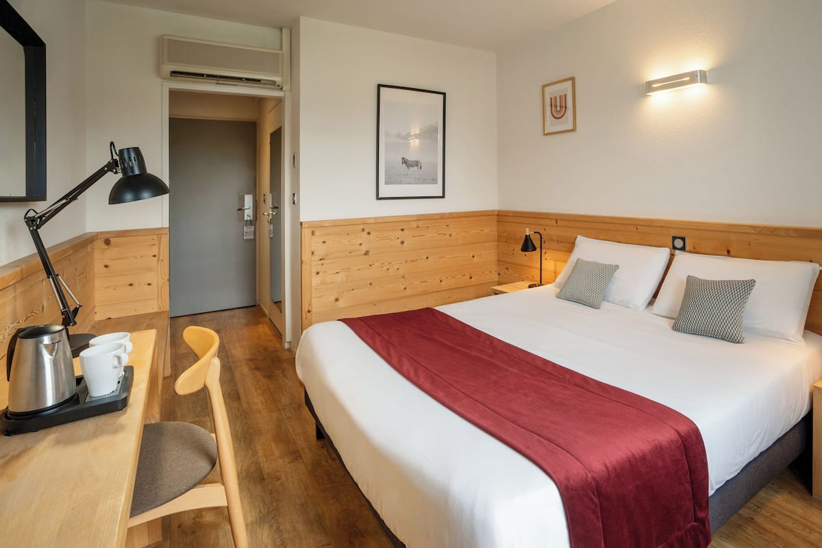 Habitación de hotel con cabecera de madera, cama grande con ropa de cama blanca y roja, mesita de noche con lámpara, silla, cafetera y un cuadro en la pared.