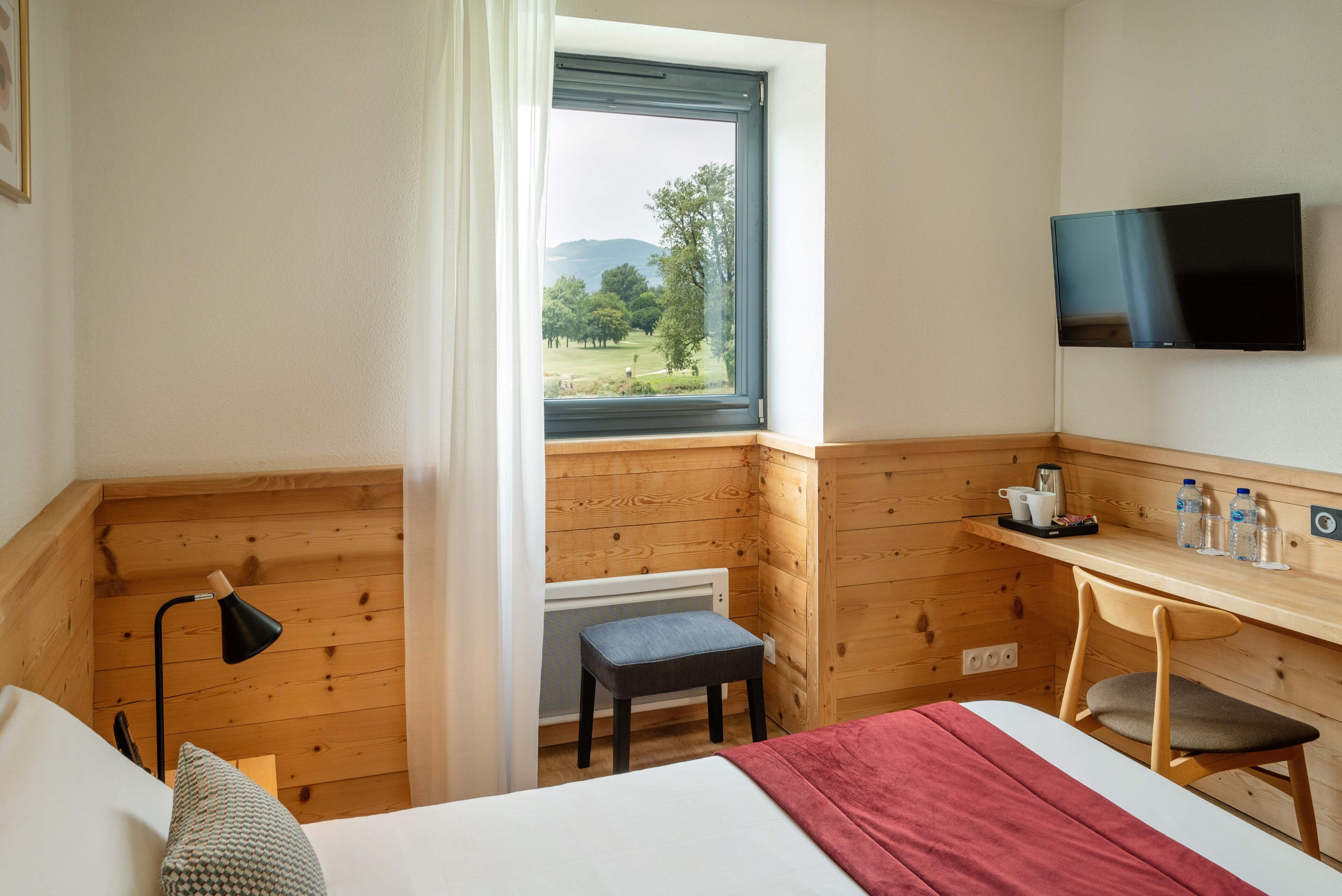 Habitación de hotel con cama, escritorio, silla, televisor, ventana y vistas a la vegetación, además de un radiador.