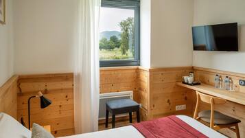 A hotel room with a bed, a desk, a chair, a TV, a window with a view of greenery, and a radiator.