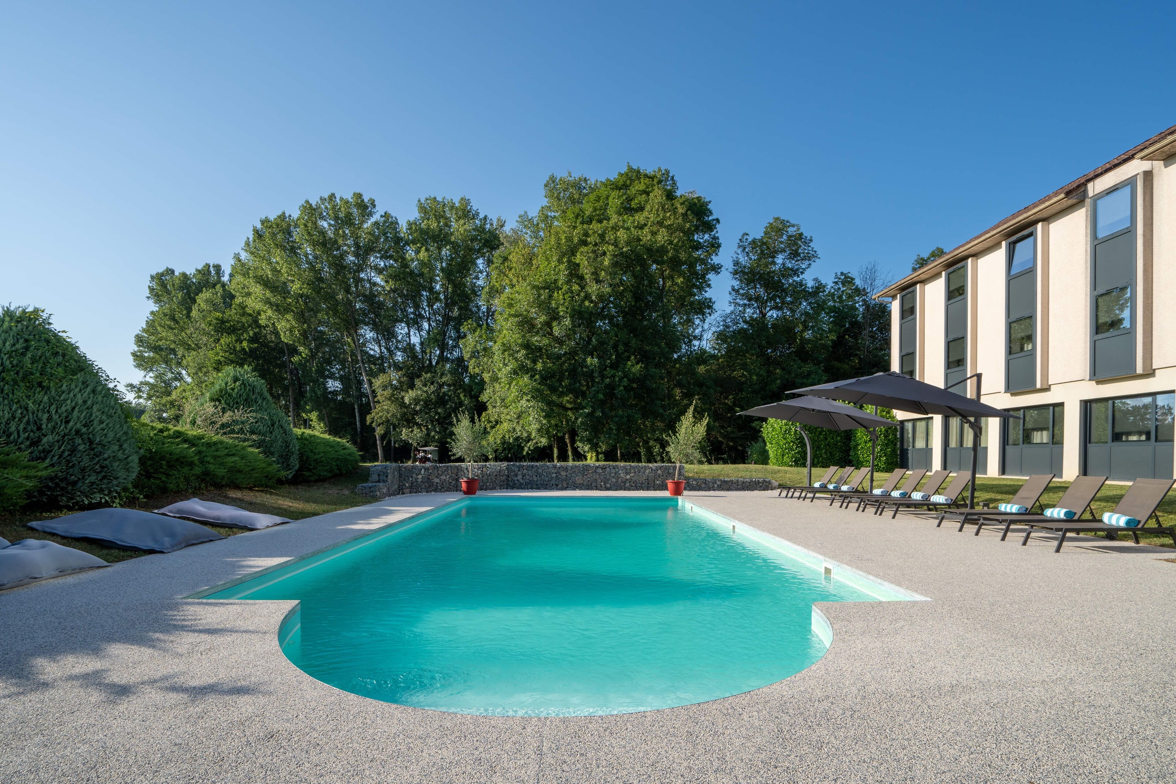 Une piscine rectangulaire dont les bords sont recouverts de petits galets, entourée de chaises longues et de parasols, avec un mur de pierre et des arbres en arrière-plan.