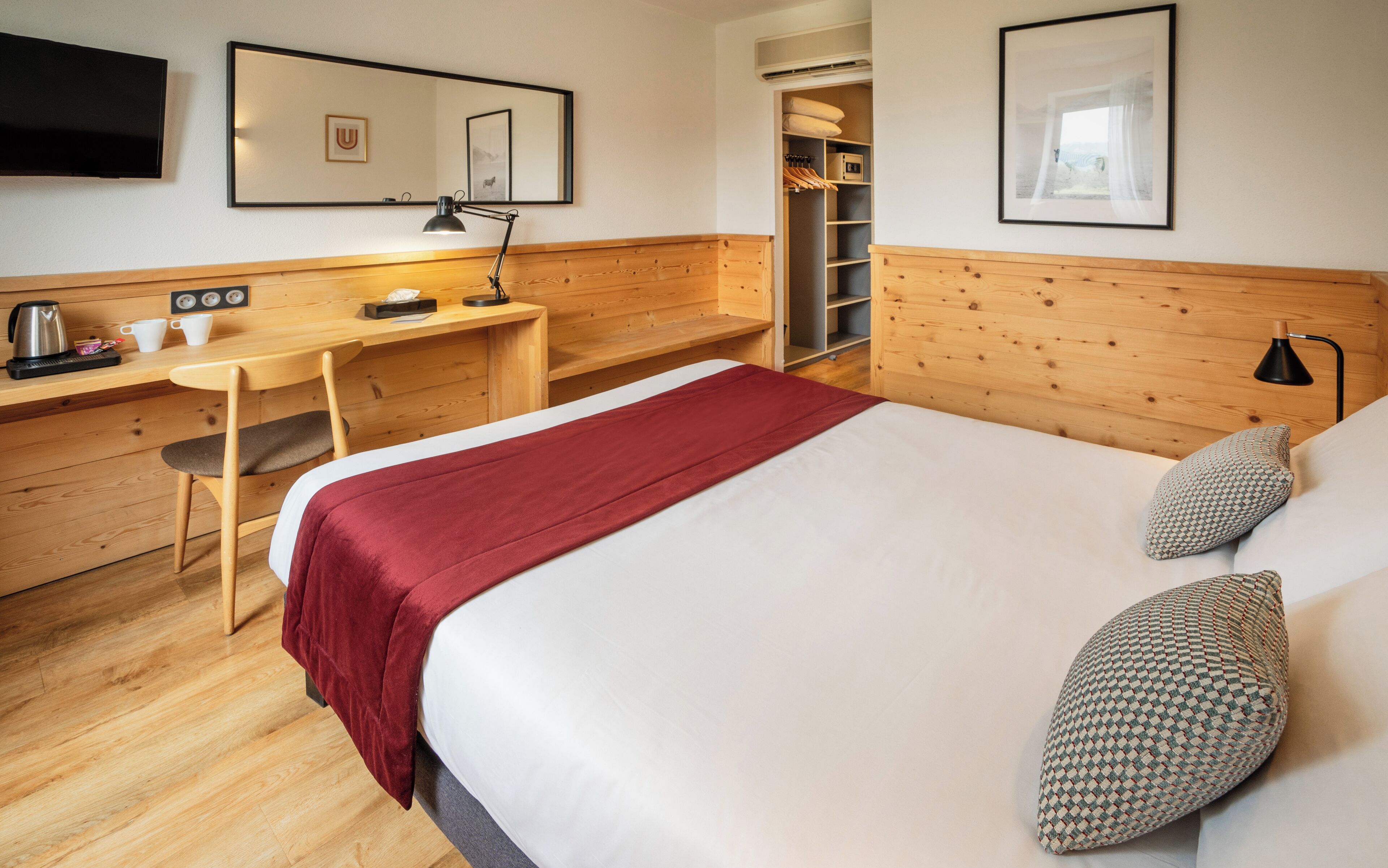 A hotel room with a wooden headboard, a bed with a red blanket, a desk with a chair, a TV, and a wardrobe.