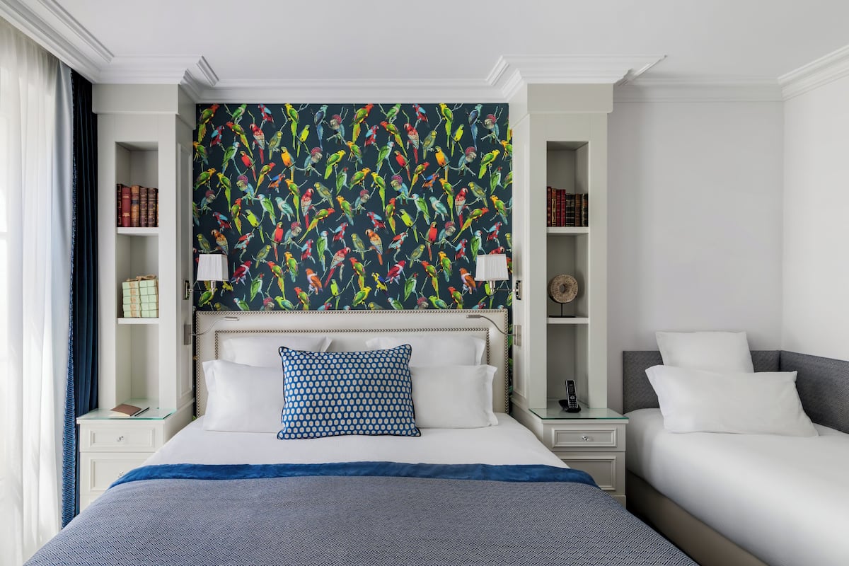 A hotel room with a large bed, a smaller bed, a bookshelf, and a wall with a colorful bird pattern.