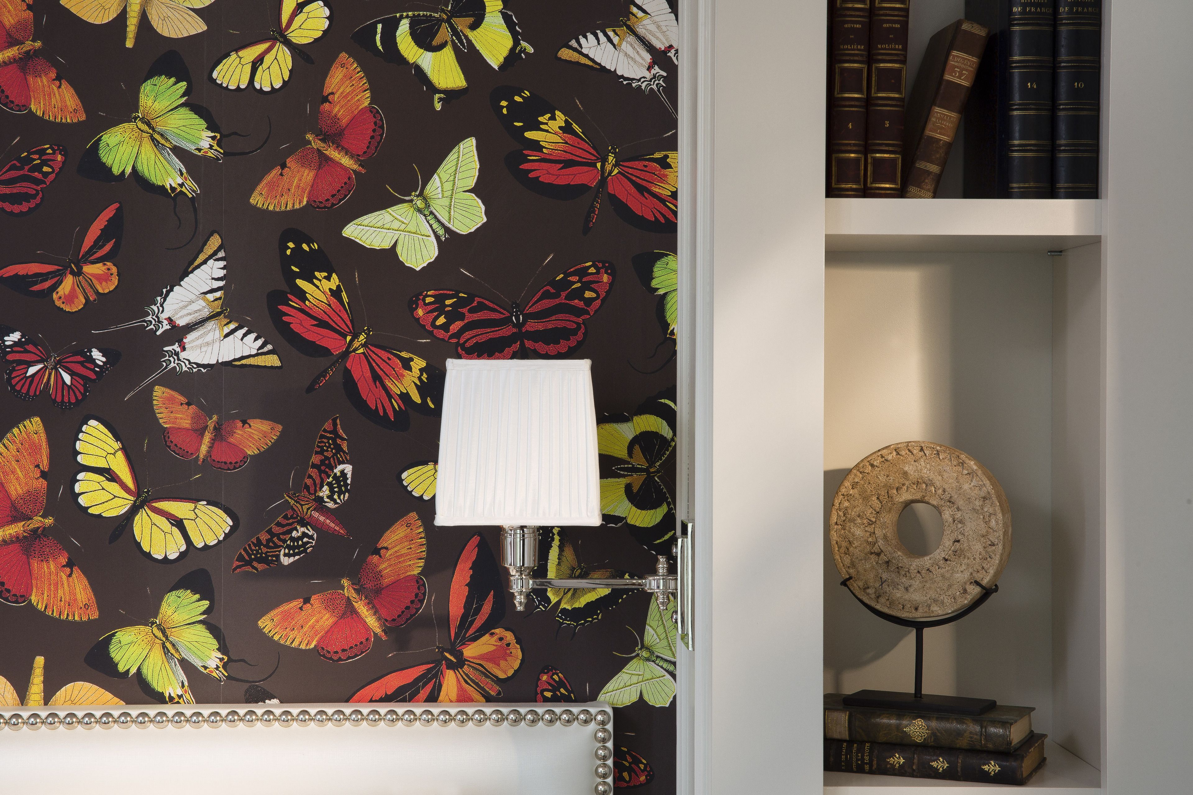 Una habitación con un papel tapiz estampado con mariposas, una lámpara blanca y un objeto decorativo redondo sobre un pedestal.