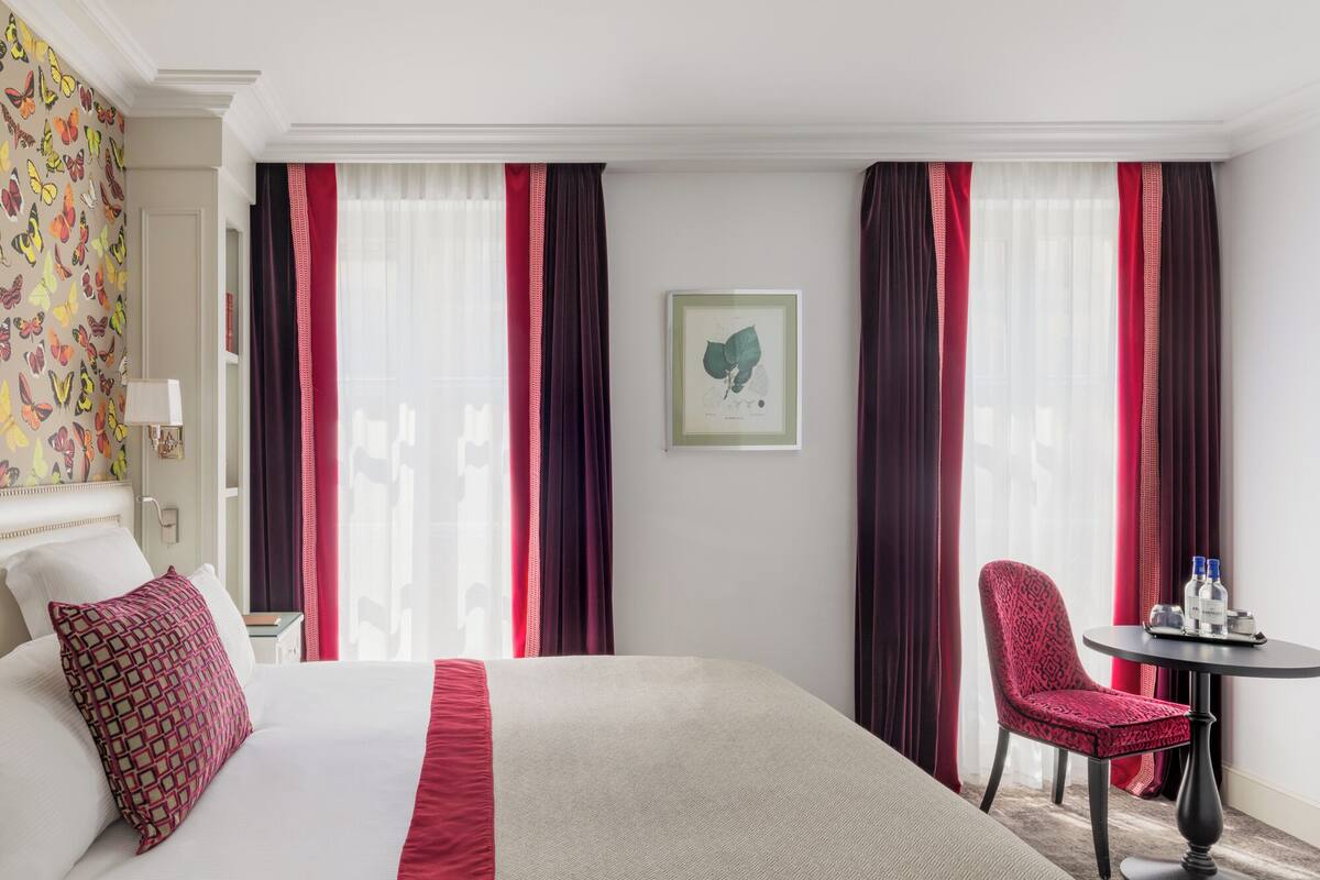 A hotel room with a large bed, a chair, a small table with bottles, and a framed botanical illustration on the wall.