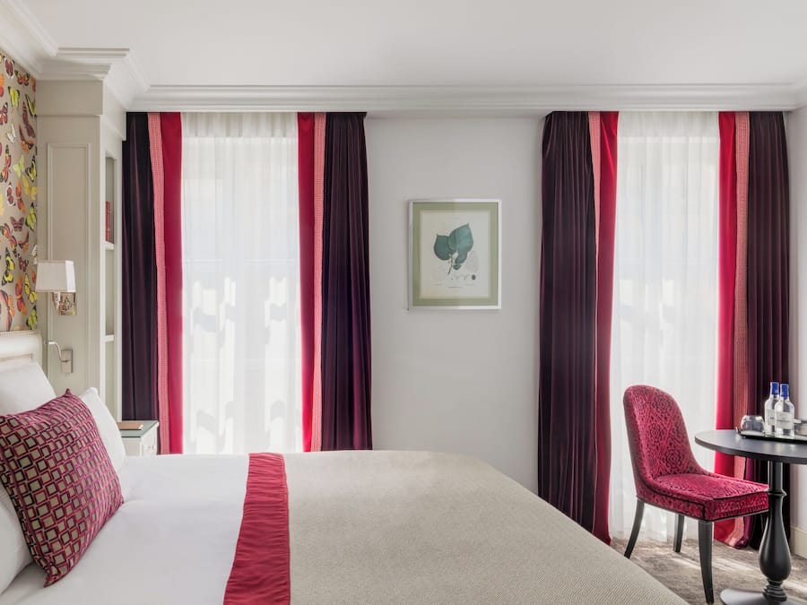 Une chambre d’hôtel avec un grand lit, une chaise, une petite table avec des bouteilles et, au mur, une illustration botanique encadrée.