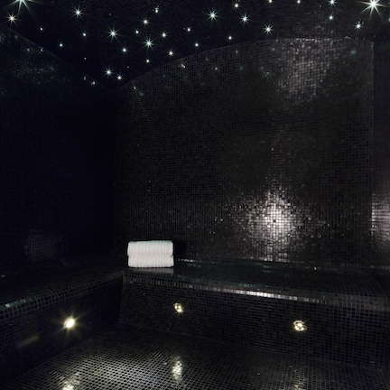 Una habitación con paredes de azulejos oscuros, una toalla blanca sobre un banco, pequeñas luces en el suelo y un techo con estrellas.