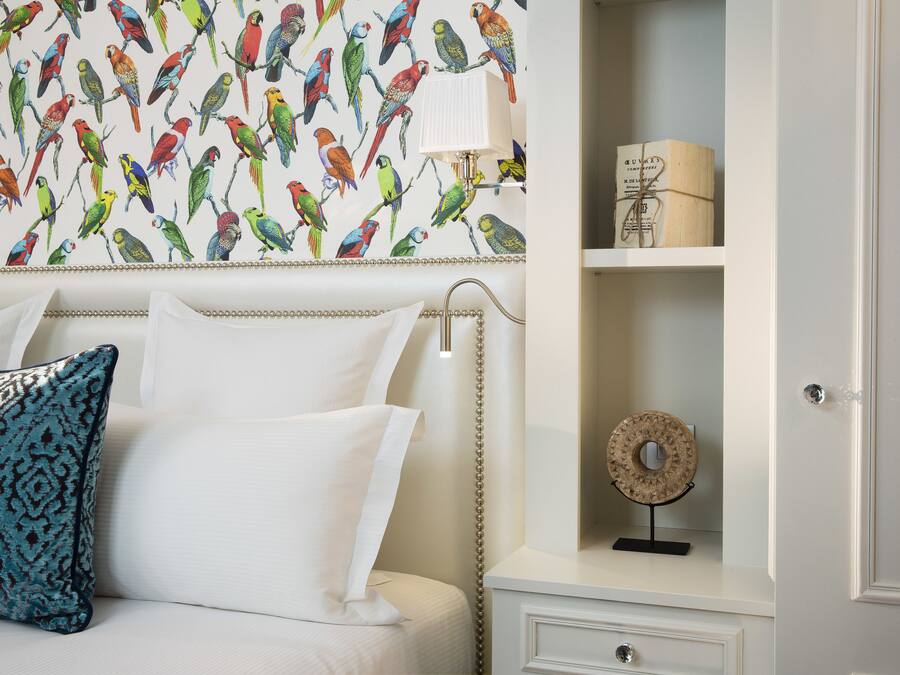 Une chambre à coucher avec un papier peint à motifs, un lit blanc agrémenté de coussins décoratifs, une table de chevet avec une lampe et une petite étagère sur laquelle est posé un objet décoratif.