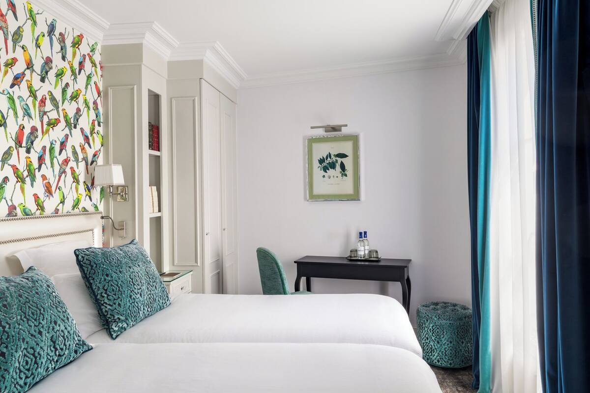 A hotel room with a large bed, a nightstand, a desk, a chair, a window with curtains, and a wall with a bird pattern.