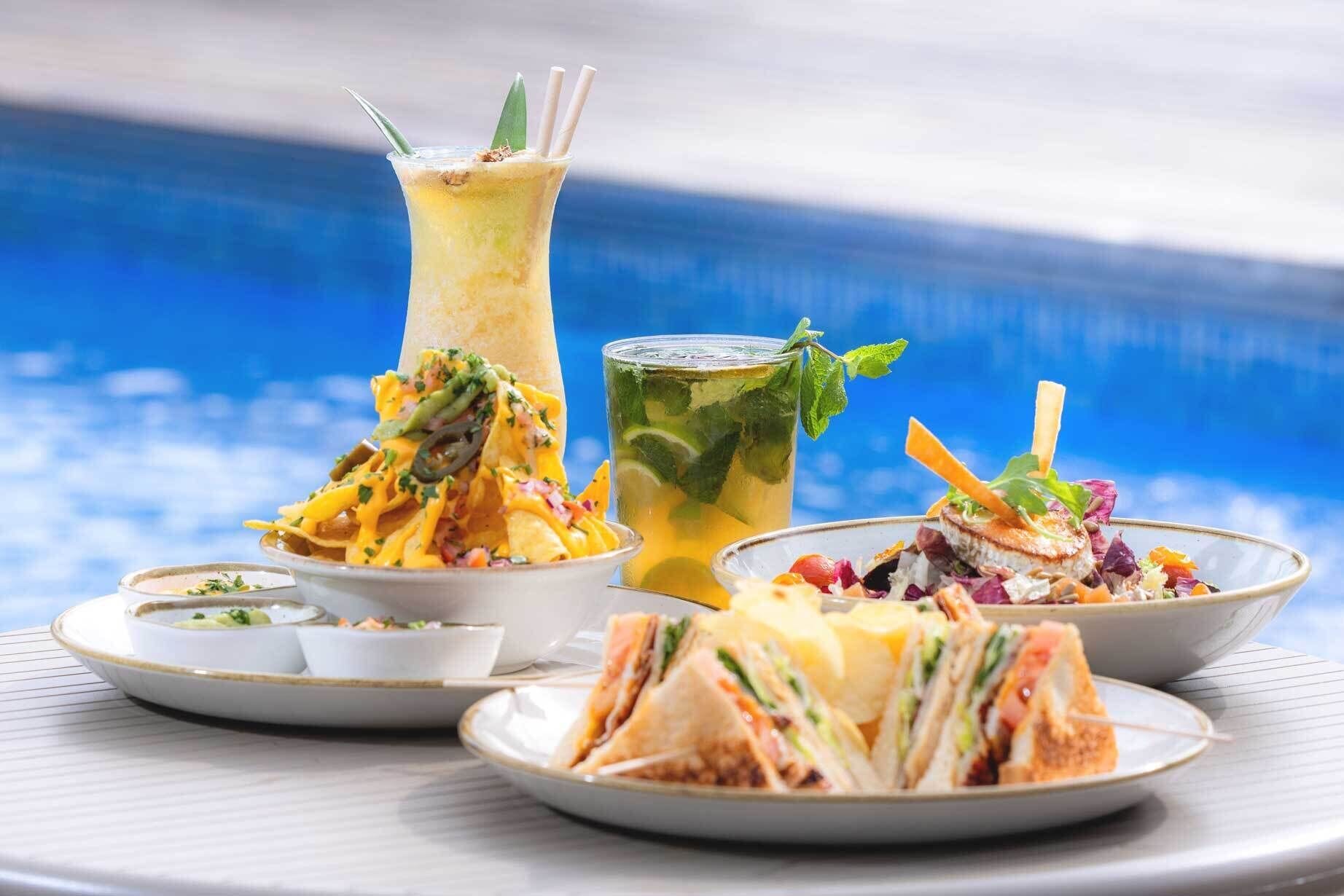 Una mesa con diferentes platos, incluyendo nachos, una ensalada y sándwiches, con una piscina al fondo.