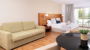 Habitación de hotel con sofá, cama con almohadas, mesita de noche con lámpara, una pequeña mesa con una planta y vistas a través de una ventana.