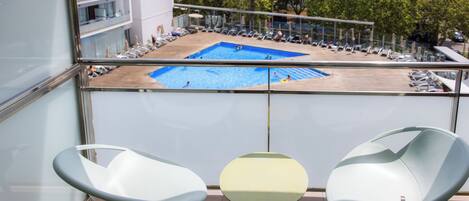 A balcony with two chairs and a small table, overlooking a swimming pool with lounge chairs and umbrellas.