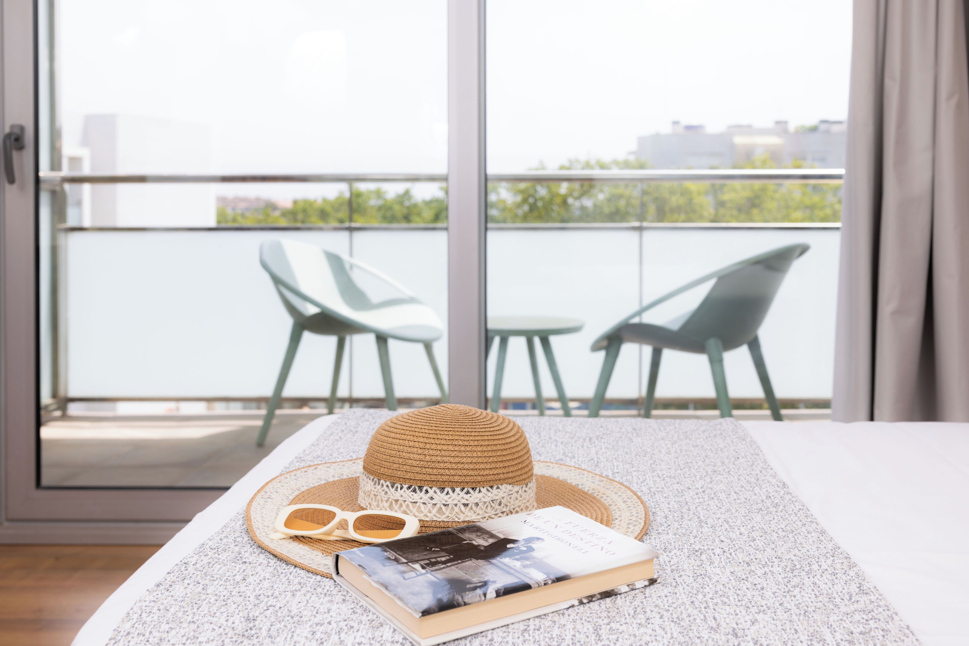 Un balcón con una mesa dispuesta, un sombrero de paja, gafas de sol y un libro.