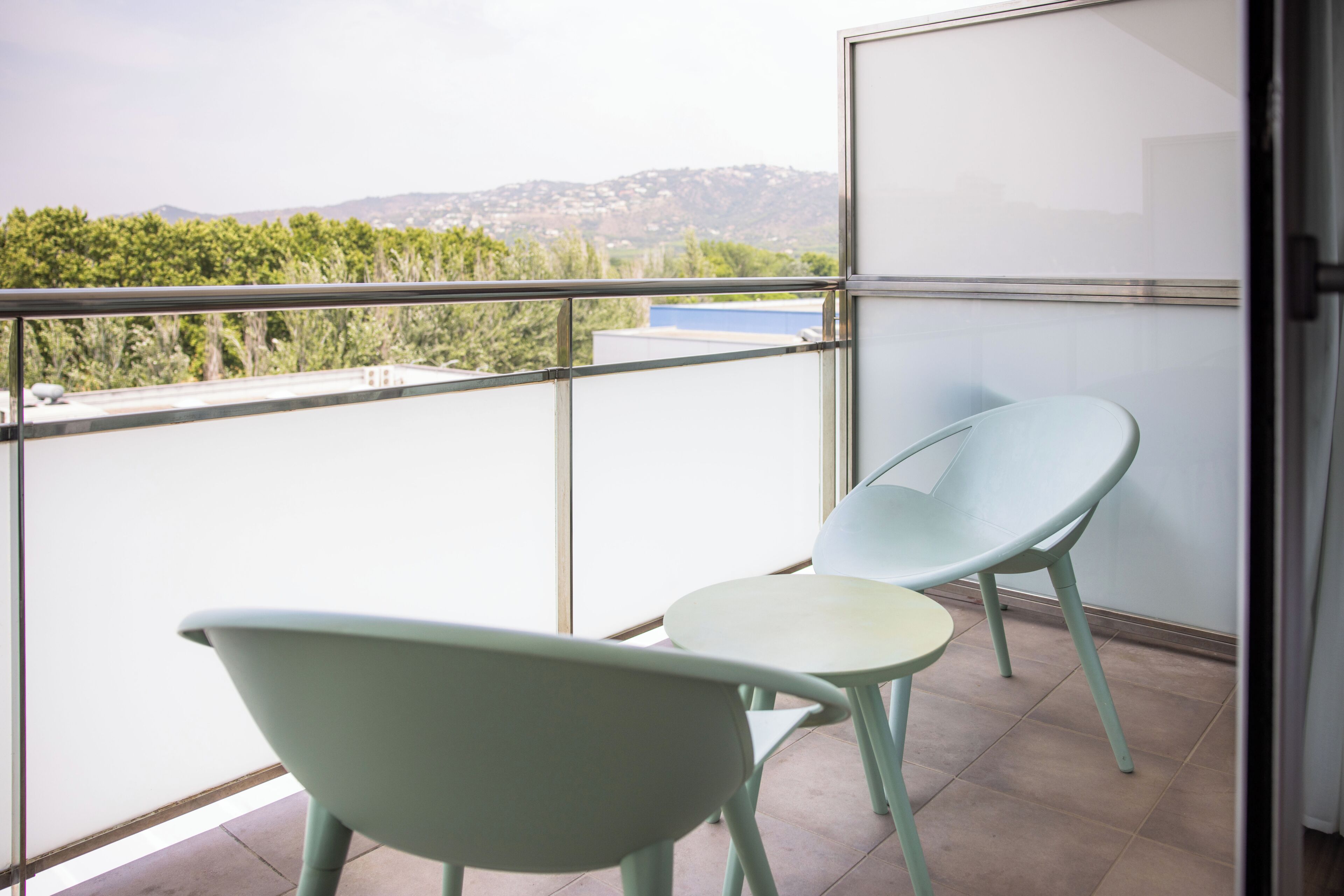 Un balcón con dos sillas, una mesa, un ventanal de vidrio y vistas a zonas verdes y colinas al lejano.