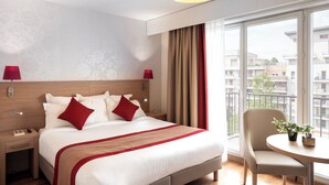 Quarto de hotel com uma cama grande, duas mesas de cabeceira, um abajur vermelho, uma mesinha com uma planta e vista para a cidade através de amplas janelas.