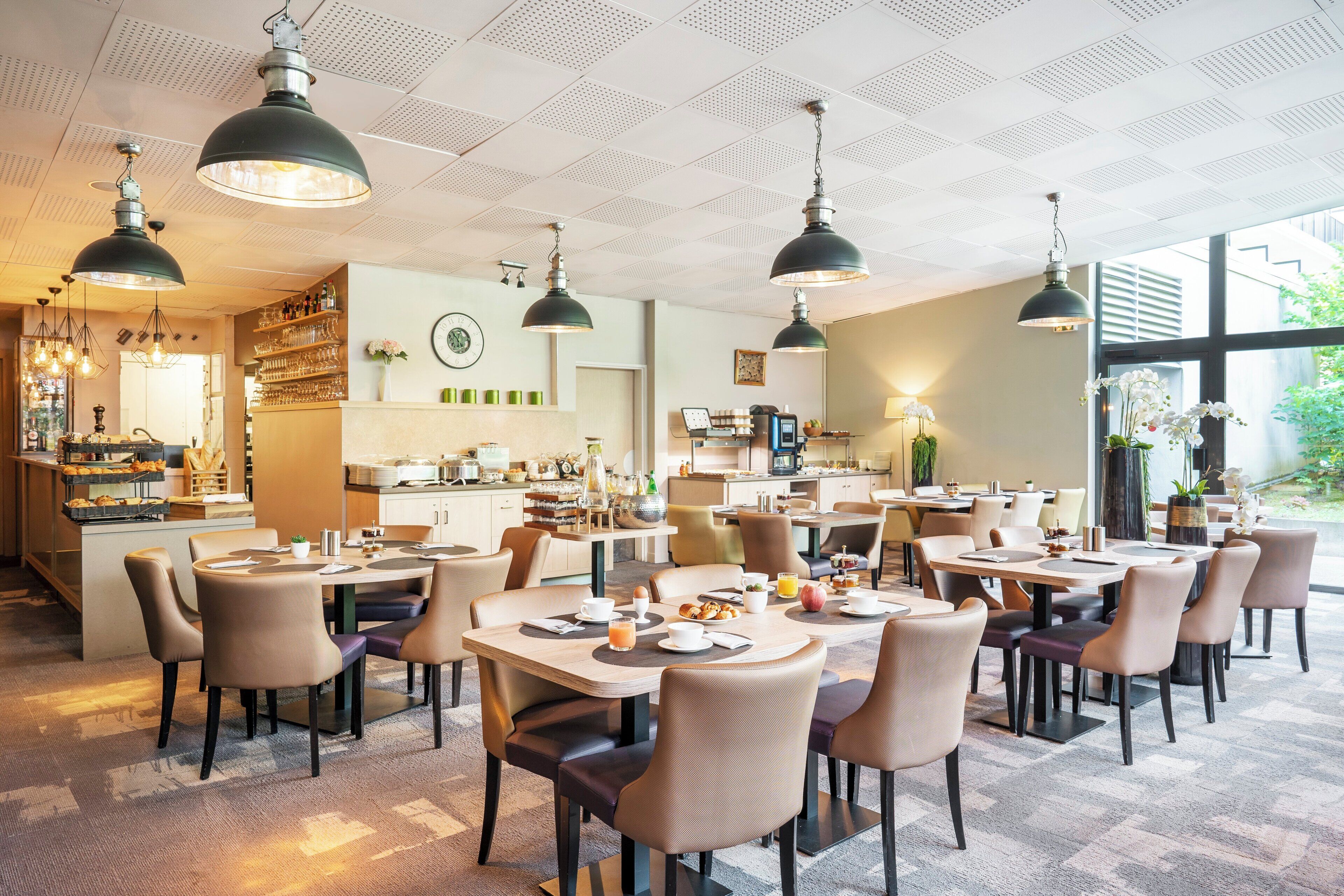 A modern hotel dining area with multiple tables and chairs, pendant lighting, and a kitchen in the background.
