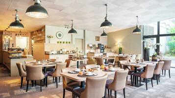 A modern hotel dining area with multiple tables and chairs, pendant lighting, and a kitchen in the background.