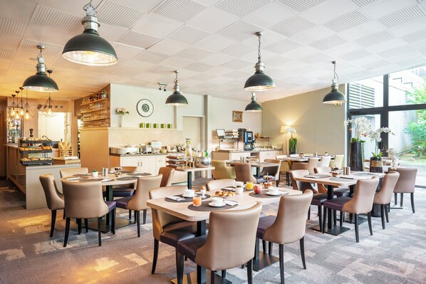 Un comedor de hotel moderno con varias mesas y sillas, iluminación colgante y una cocina al fondo.
