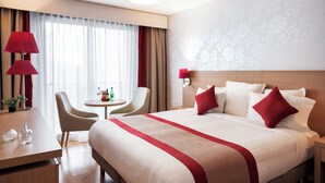 Quarto de hotel com uma cama grande, uma escrivaninha, uma cadeira, uma mesinha e um abajur.