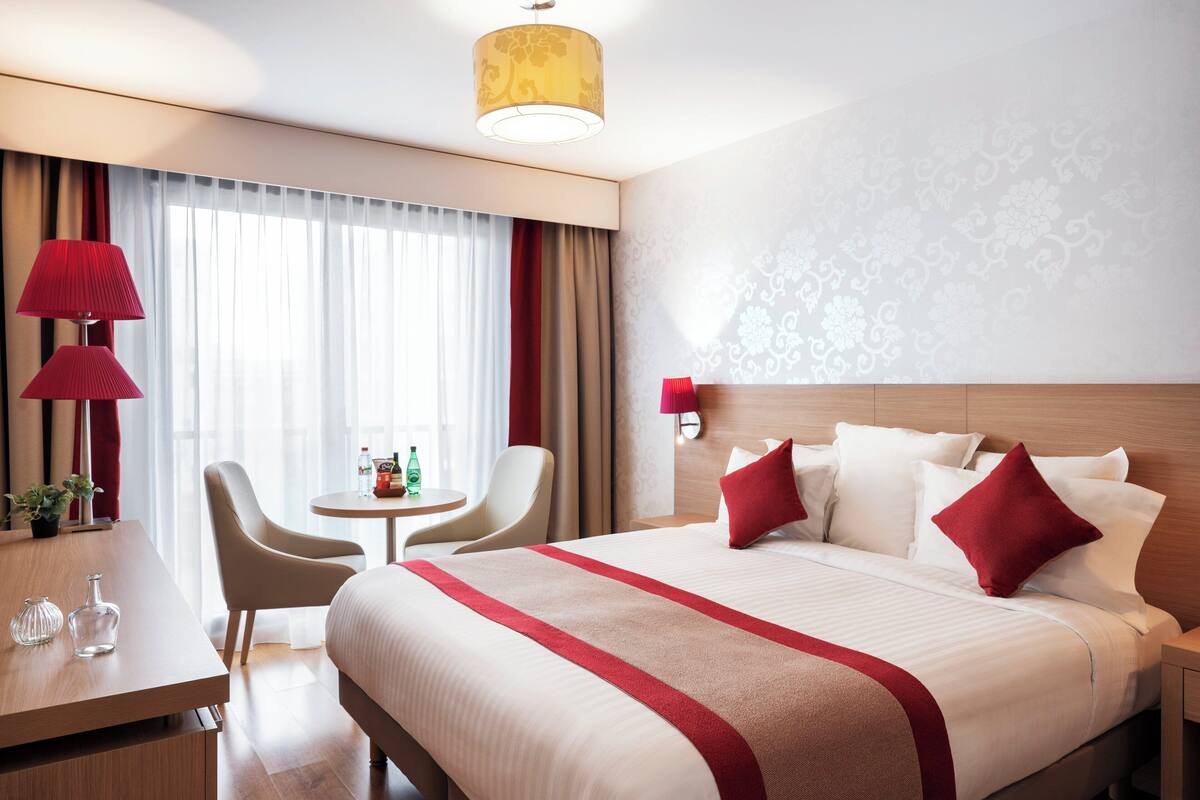 A hotel room with a large bed, a desk, a chair, a small table, and a lamp.