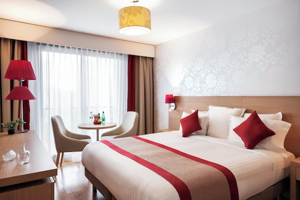 A hotel room with a large bed, a desk, a chair, a small table, and a lamp.