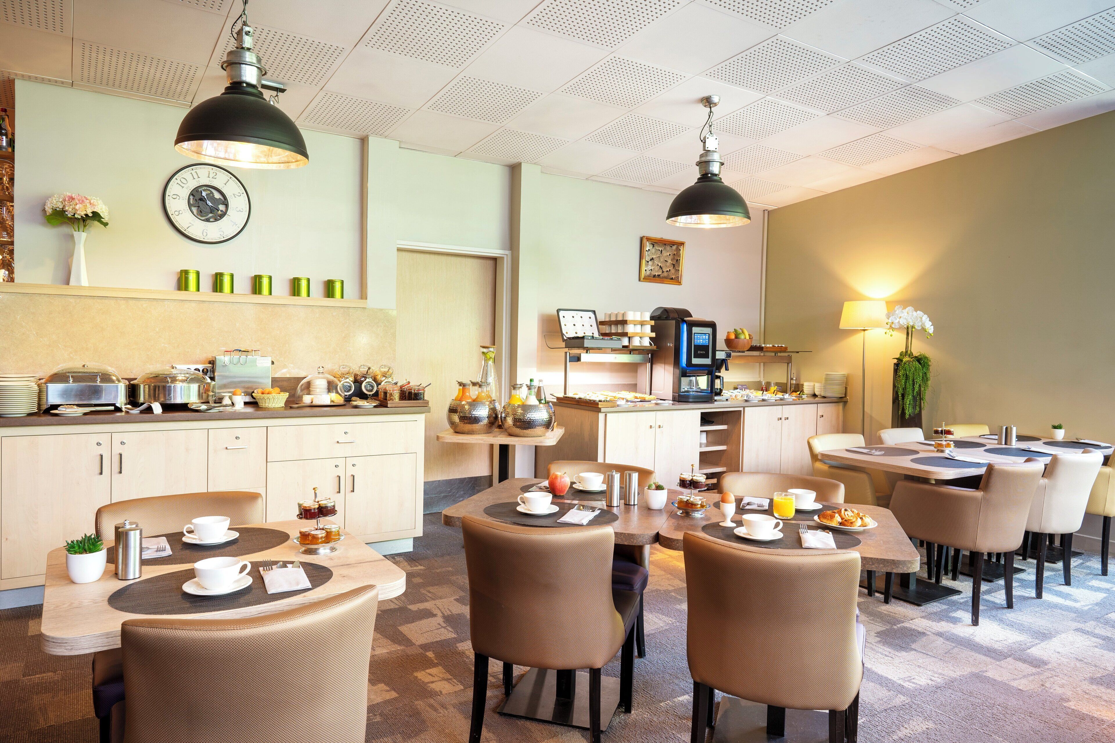 A hotel dining area with multiple tables and chairs, a kitchen in the background, and a clock on the wall.