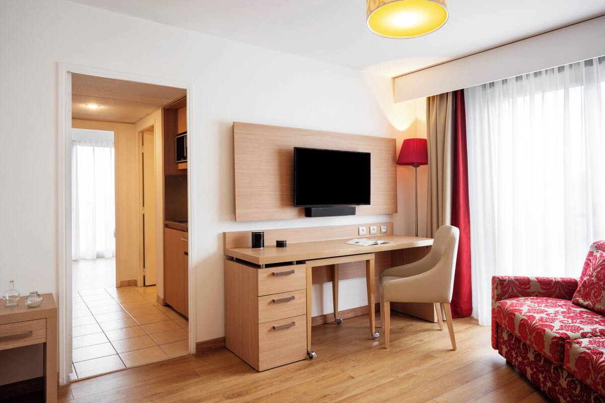 A hotel room with a wooden desk, a chair, a TV mounted on the wall, a red lamp, and a red patterned sofa.