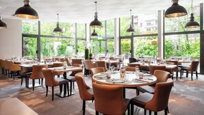 Uma sala de jantar ampla com mesas e cadeiras redondas, grandes janelas com vista para o verde e iluminação pendente.