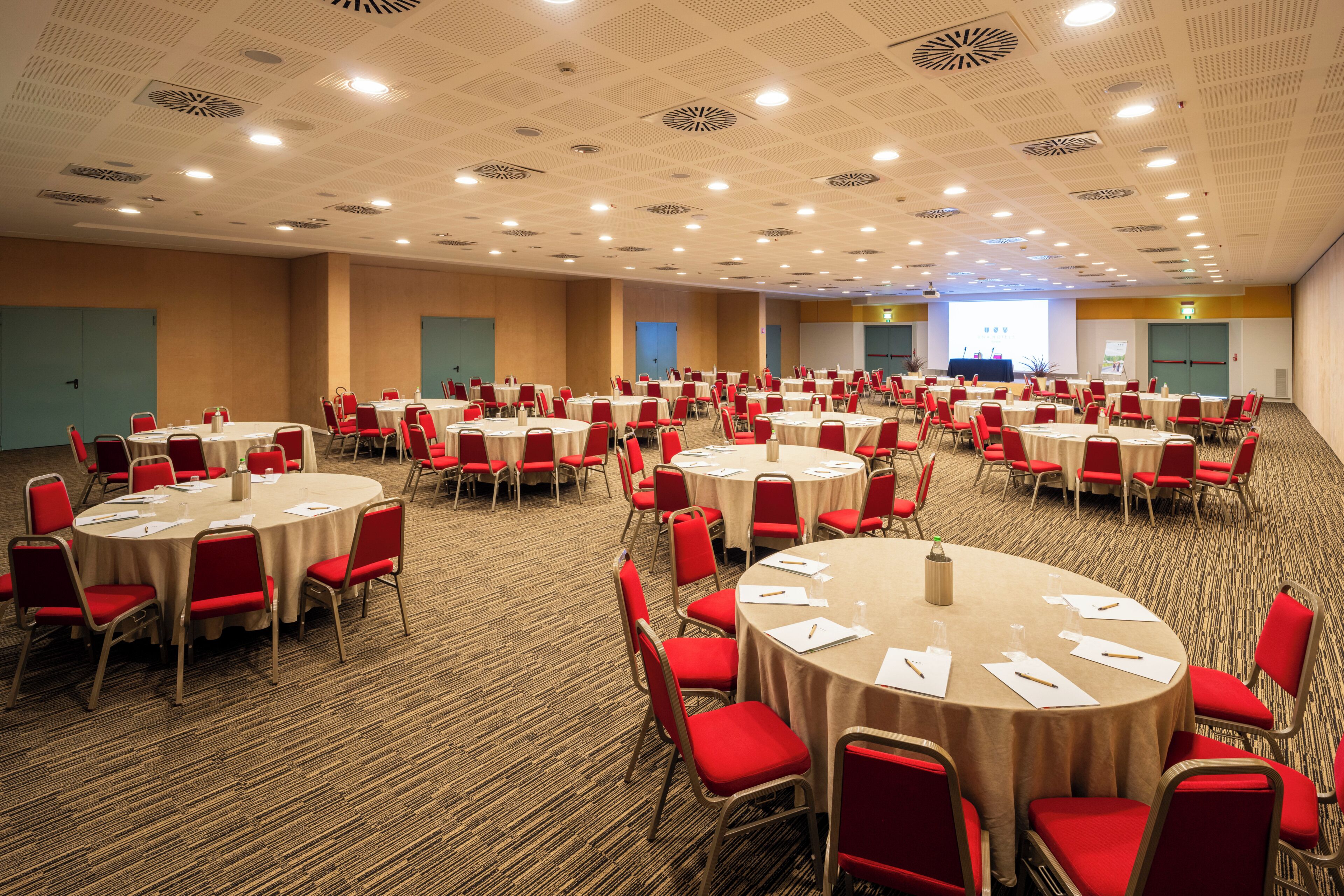 A conference room with multiple round tables, red chairs, and a carpeted floor.
