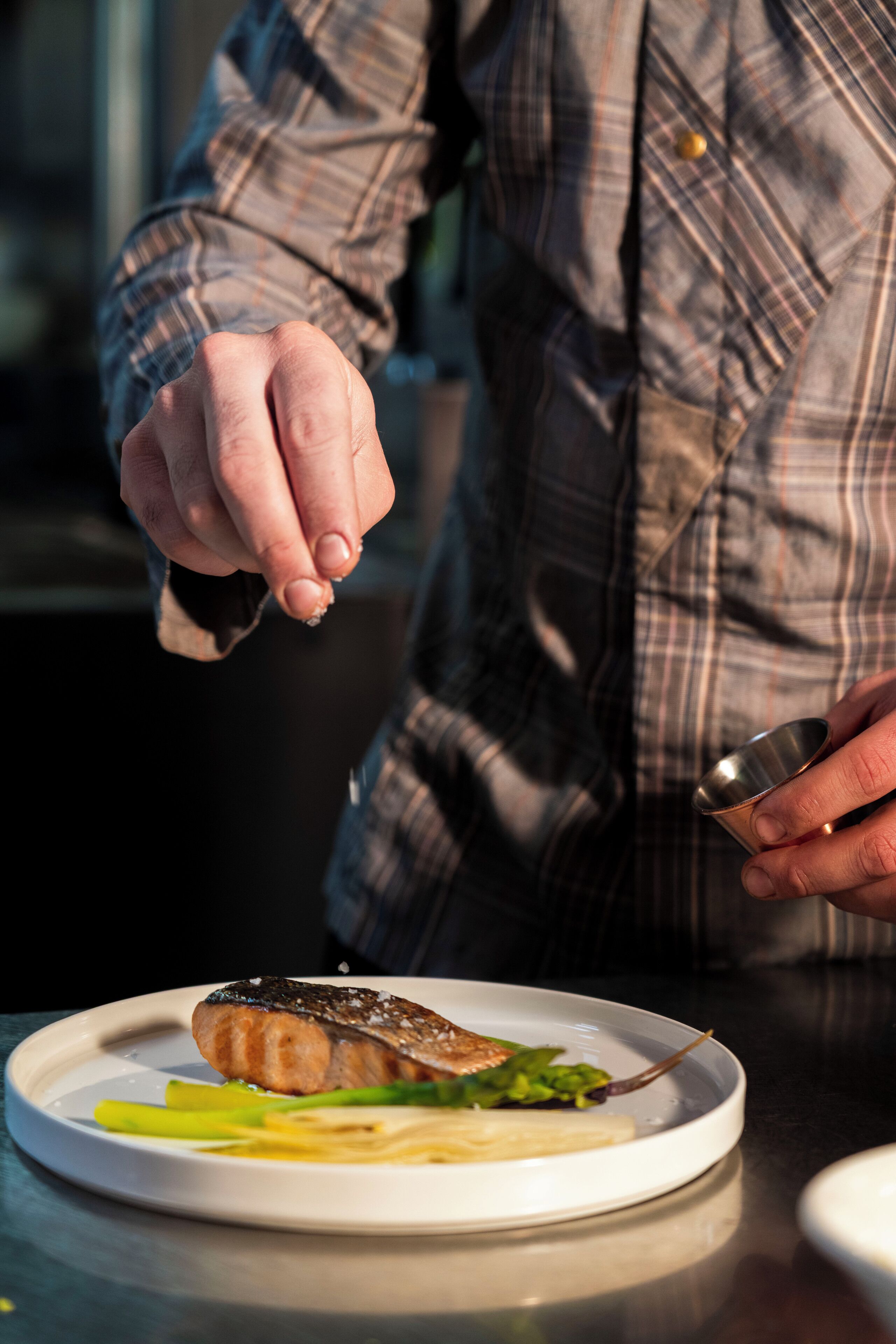 A person in a plaid shirt holding a spoon over a plate with a piece of salmon and vegetables.