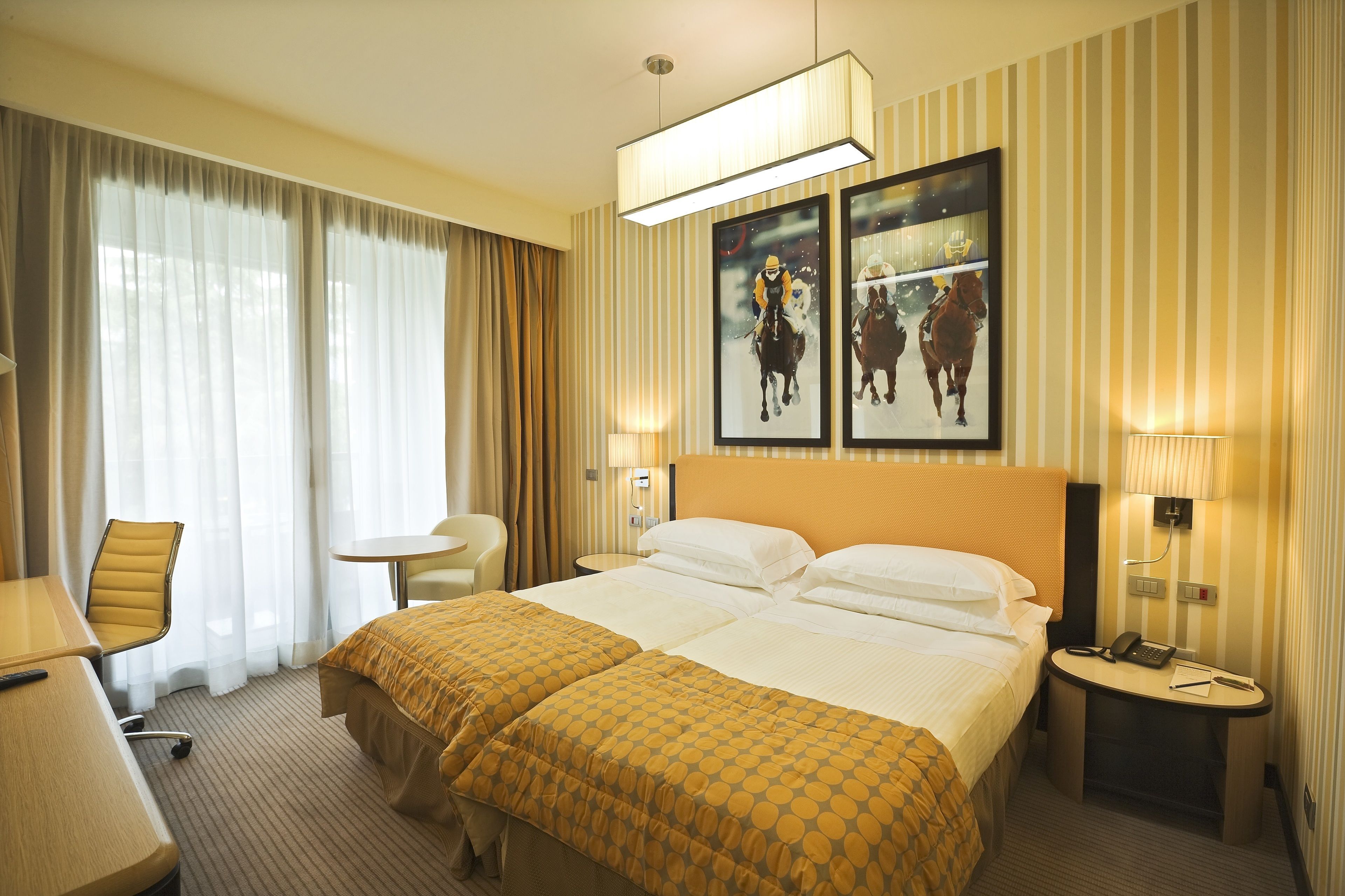 A hotel room with a large bed, a desk, a chair, and a small table. There are two framed pictures on the wall.