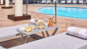 Un área junto a la piscina con sillones de descanso, una mesa con aperitivos y bebidas, y una piscina.