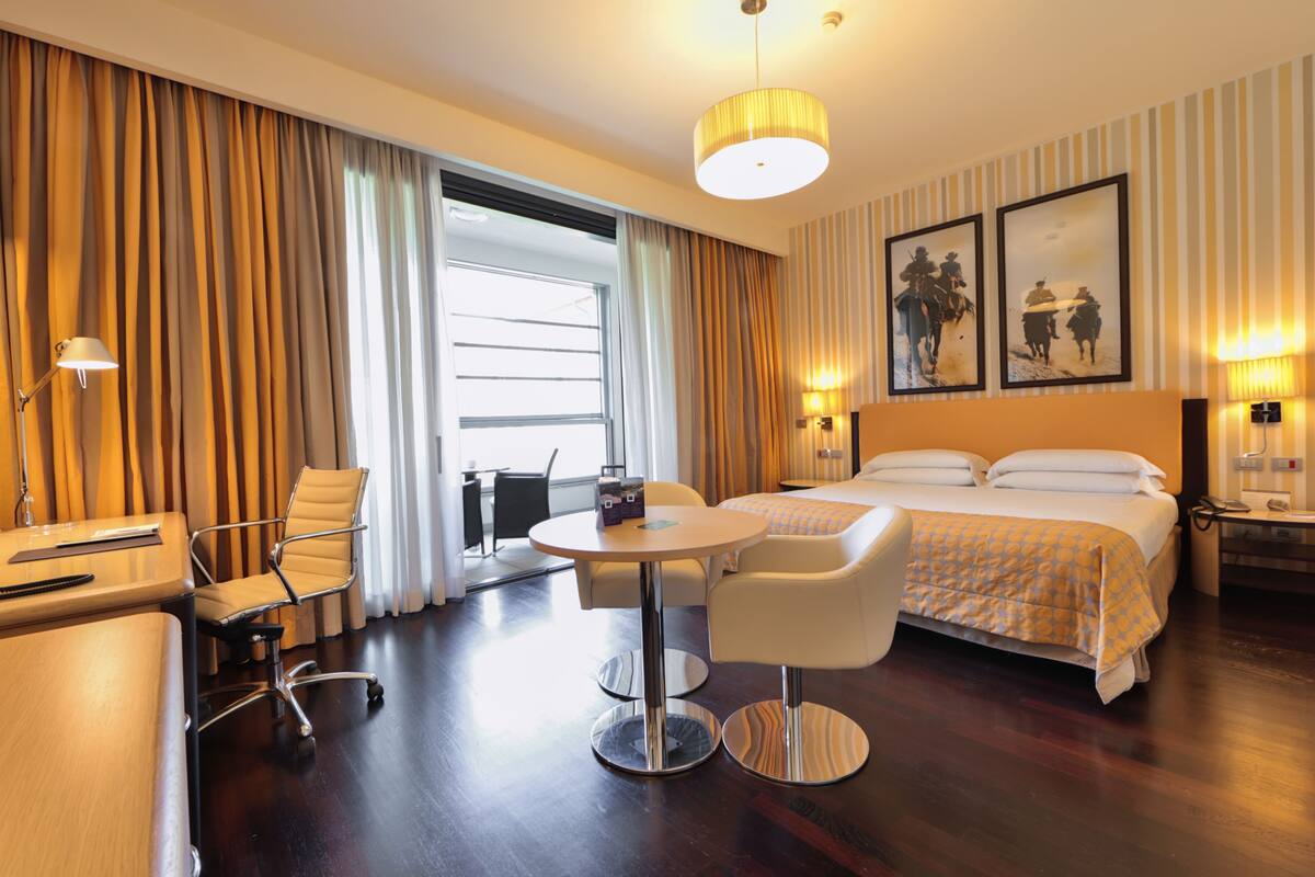 A hotel room with a bed, two chairs, a desk, a lamp, and a large window with curtains.