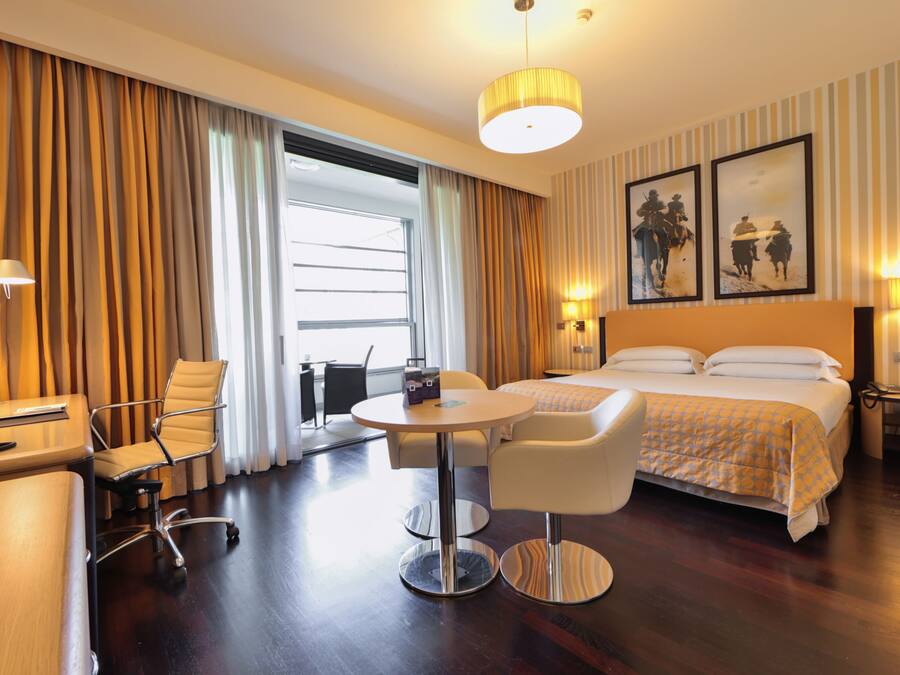 A hotel room with a bed, two chairs, a desk, a lamp, and a large window with curtains.