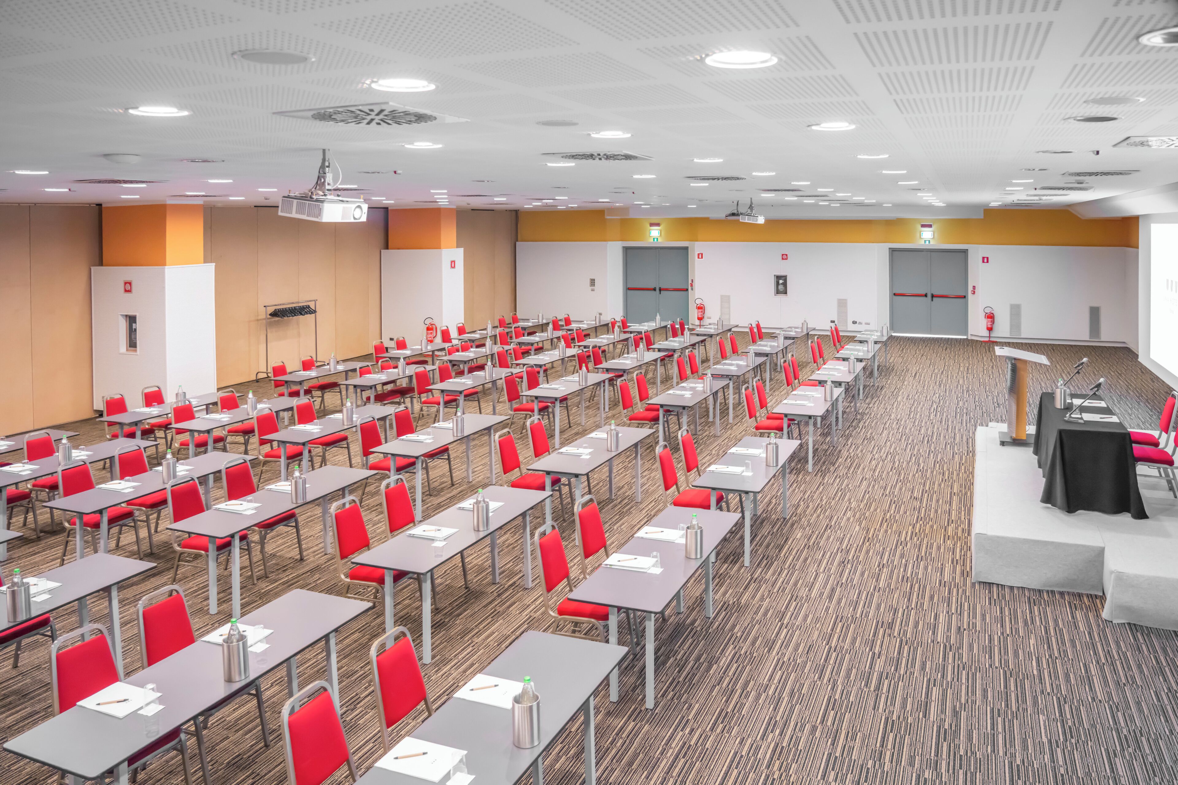 A conference room with rows of tables and chairs, a projector, and a stage with a podium.