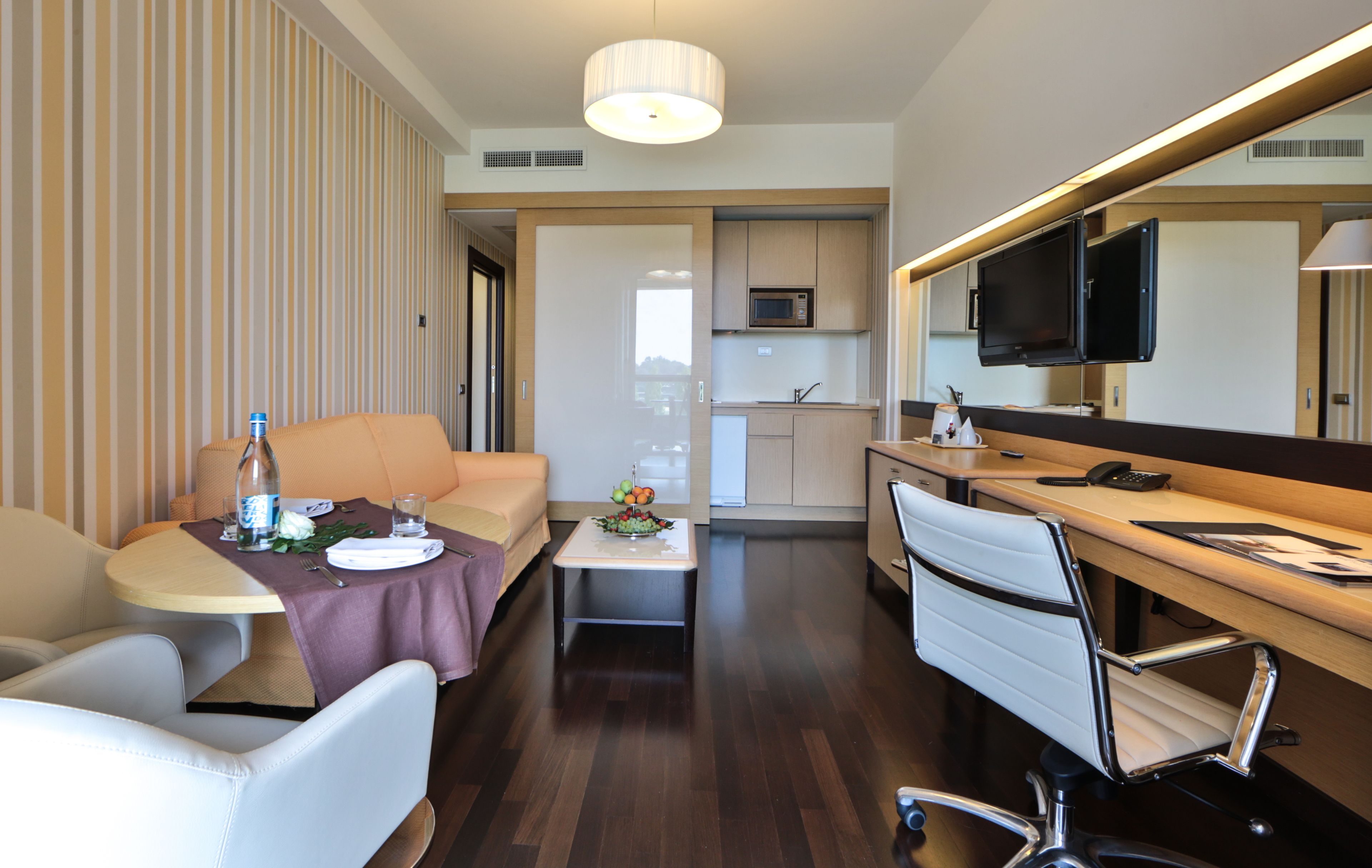 A hotel room with a dining area, a sofa, a desk, a chair, a TV, and a kitchen area.