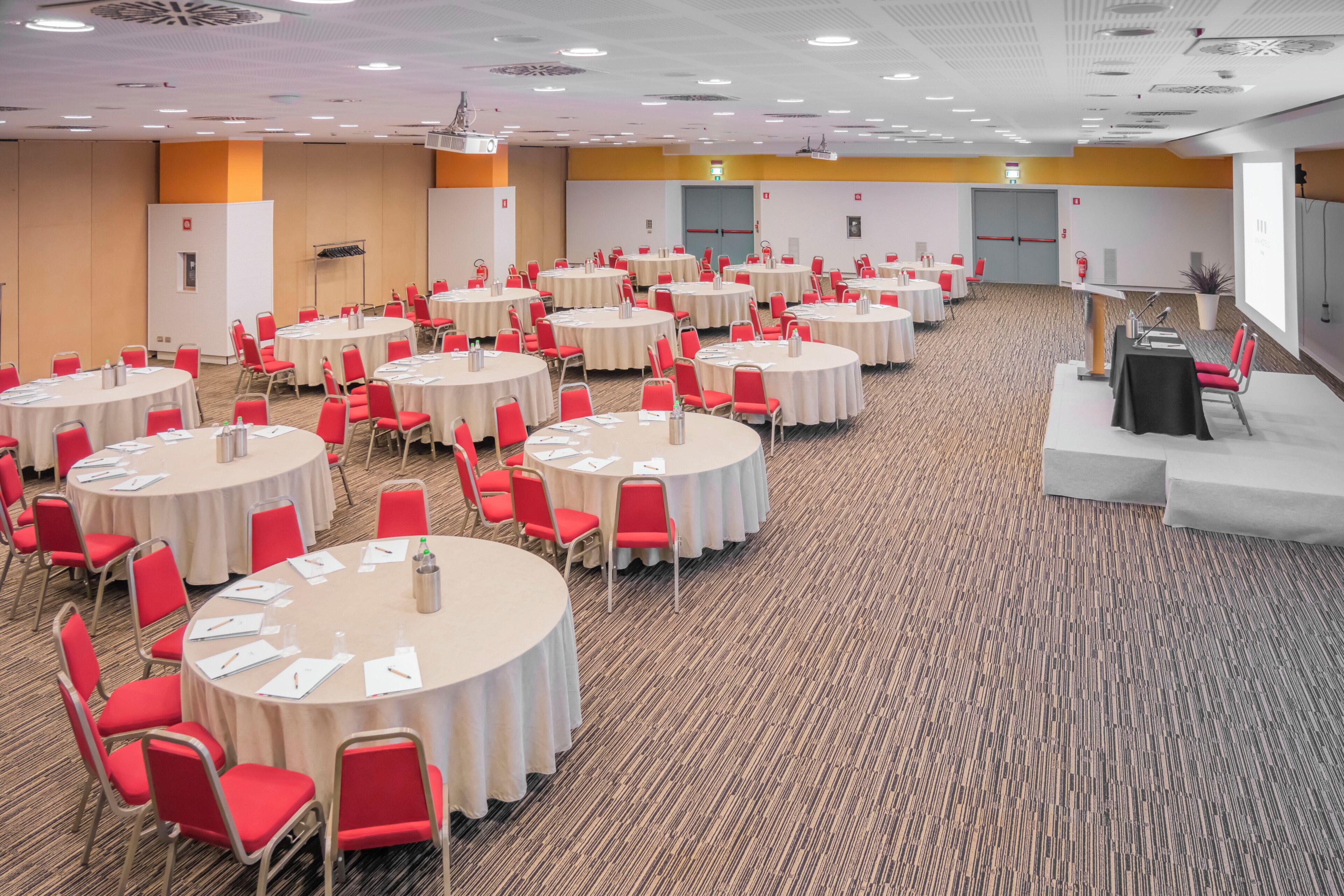Une salle de conférence dotée de plusieurs tables rondes, de chaises rouges et d’une estrade.