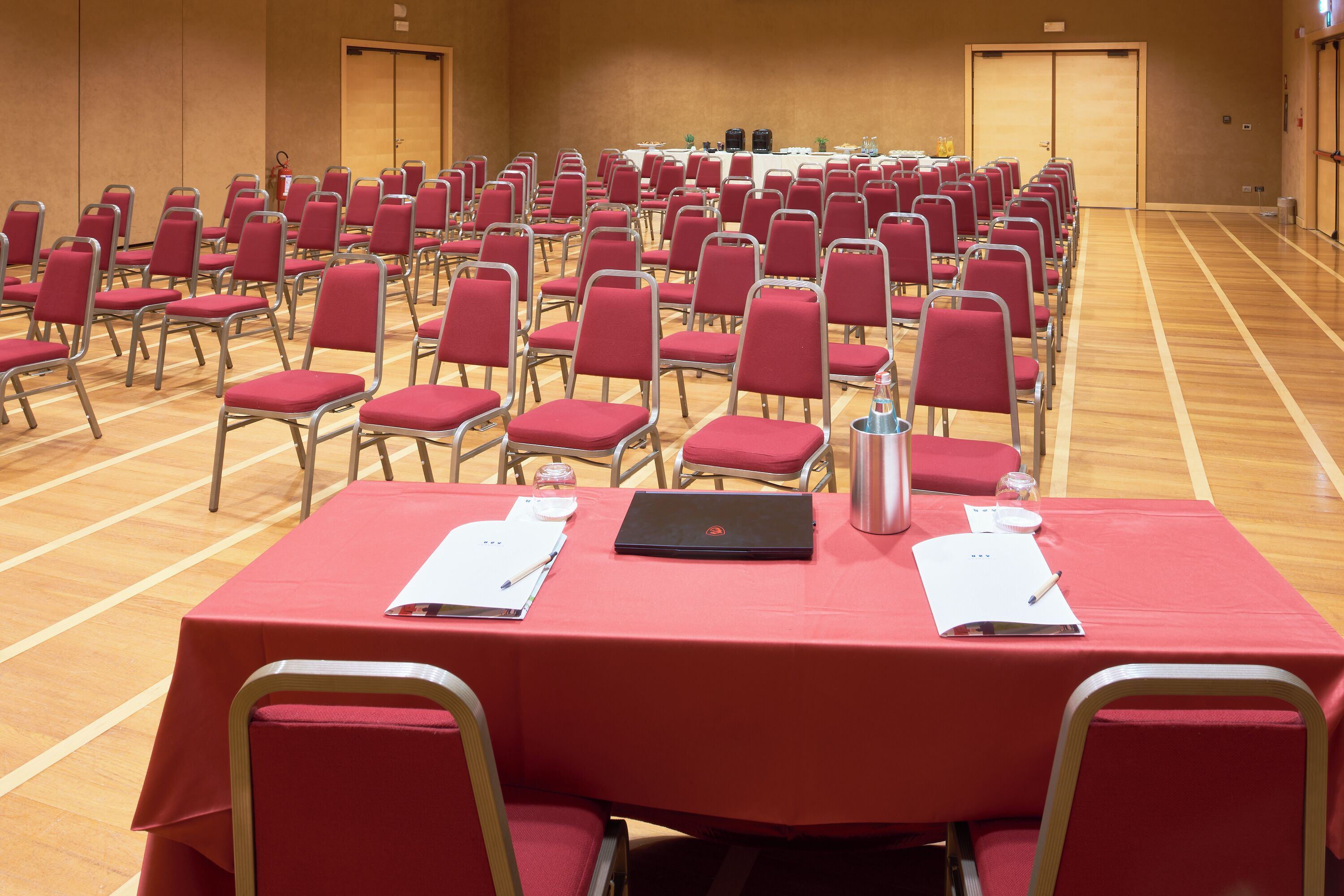 A conference room with rows of red chairs, a table with a laptop, papers, and a water bottle.