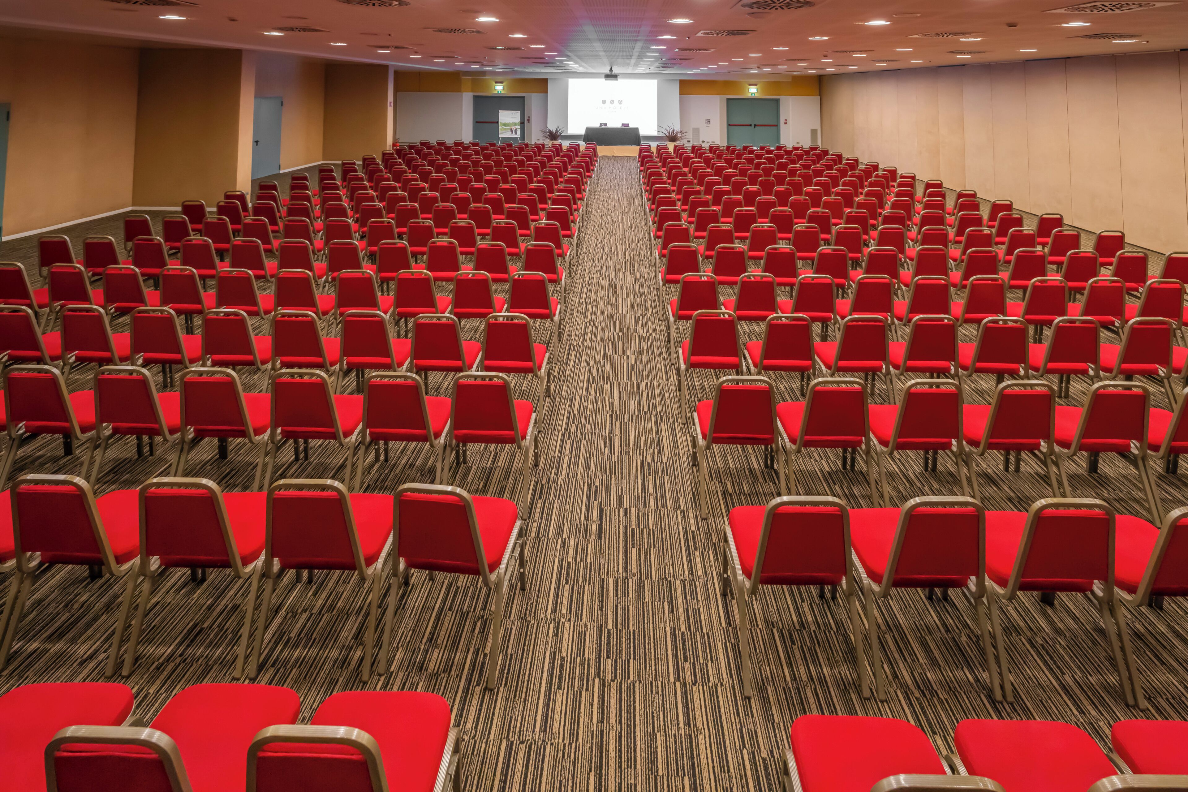 A conference room with rows of red chairs, carpeted floor, and a stage at the front.