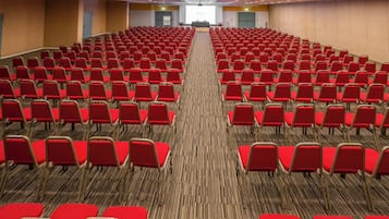 Un salón de conferencias con filas de sillas rojas, piso alfombrado y un escenario al frente.