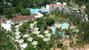 Vista aérea de un complejo turístico con varias cabañas, un área de piscina central y una playa con palmeras.
