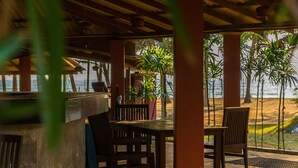 Un área de estar al aire libre con mesas y sillas de madera, un banco de hormigón y vistas a la playa y palmeras.