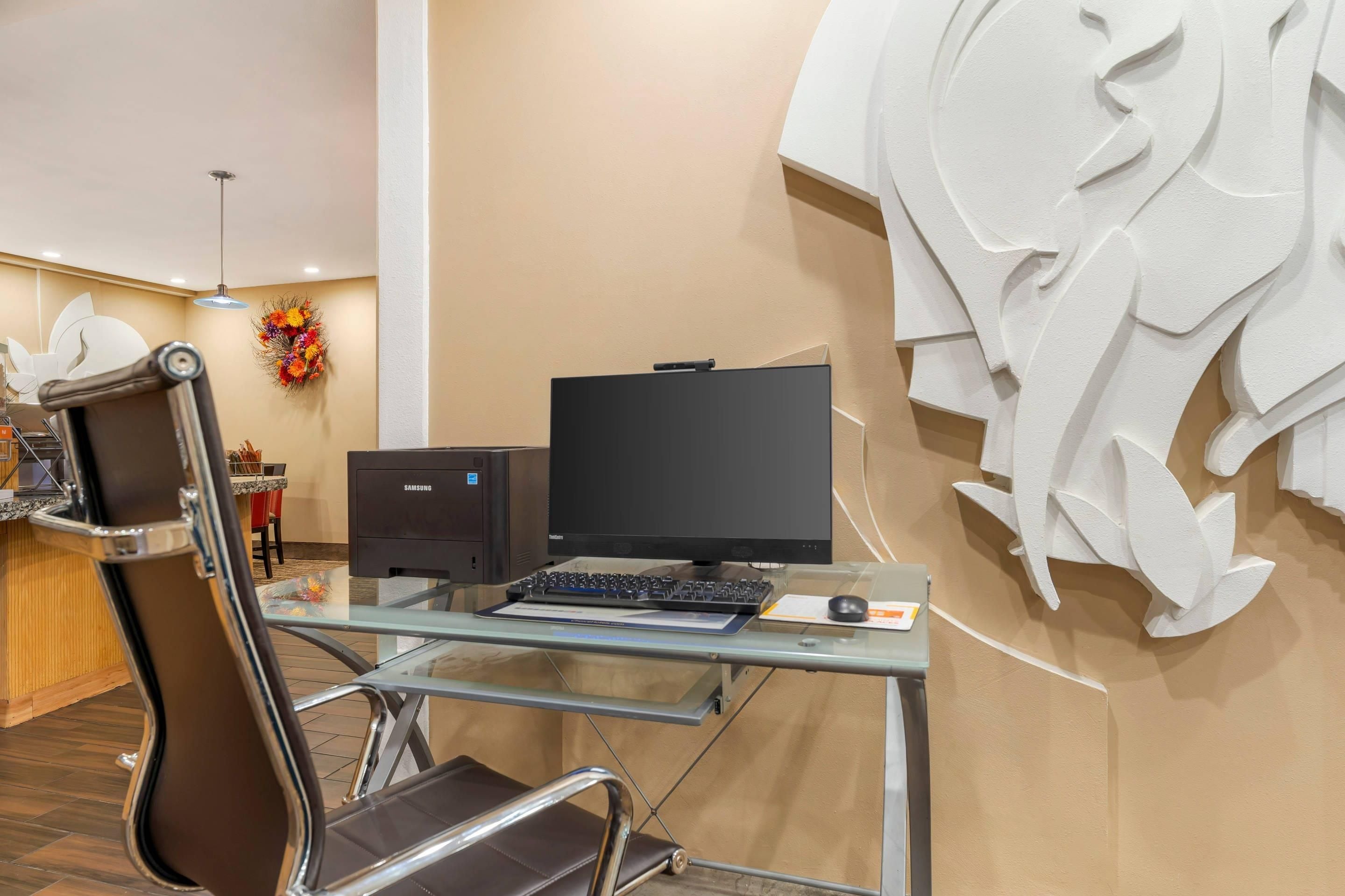 Un espace de bureau moderne avec un bureau en verre, un ordinateur noir, un fauteuil en cuir marron et un élément décoratif mural.