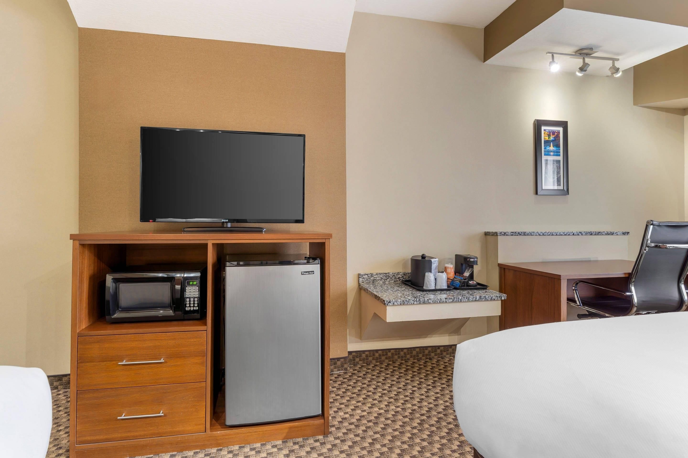 Habitación de hotel con cama, televisión, microondas, refrigerador, escritorio y cafetera, además de una silla.
