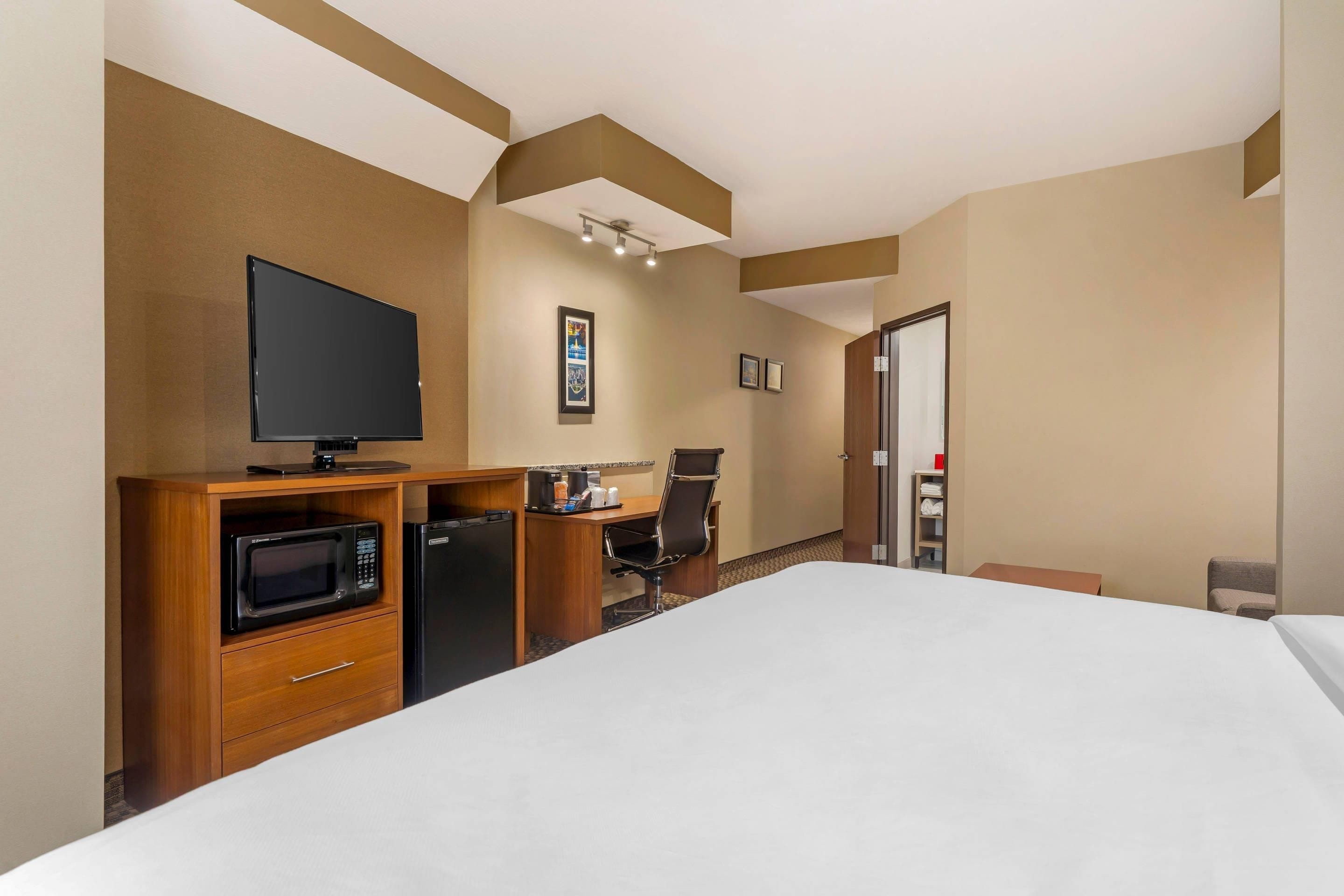 A hotel room with a bed, a television, a microwave, a refrigerator, a desk with a chair, and a door leading to another room.