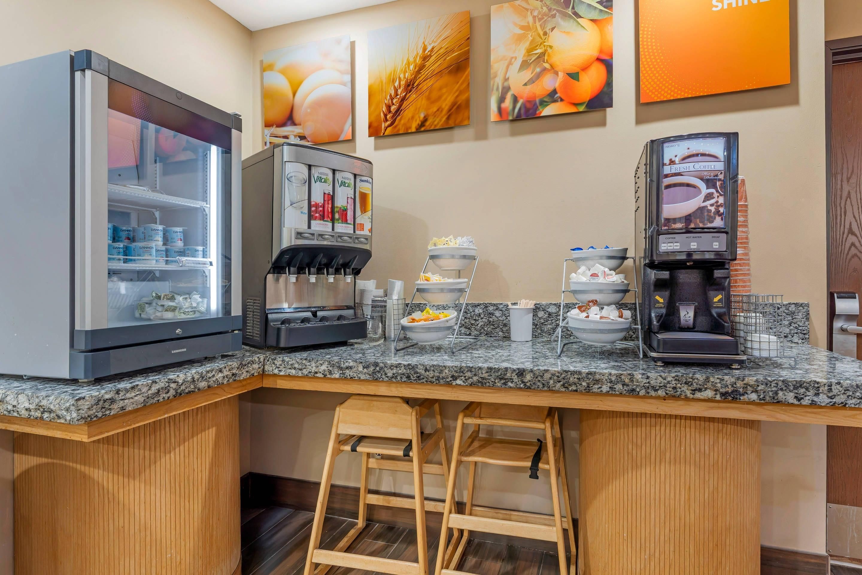 A self-service coffee station with multiple beverage dispensers, a refrigerated display case, and a counter with various food items.