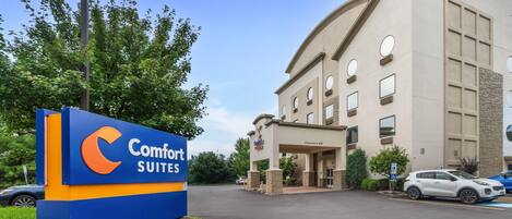 Entrada de hotel com placa da Comfort Suites, estacionamento e um edifício com várias janelas.