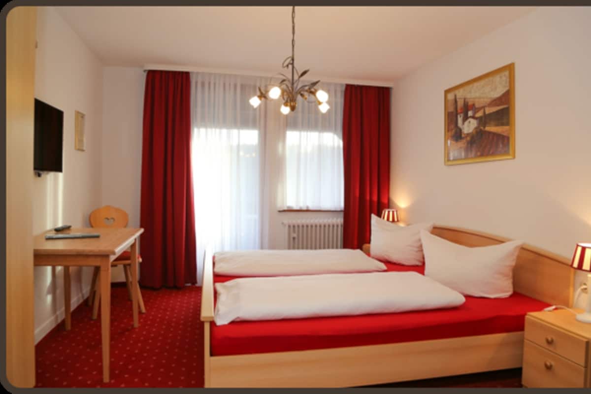 Una habitación de hotel con cama, escritorio, silla, mesita de noche, lámpara y un cuadro en la pared.