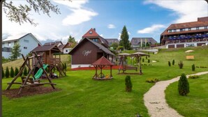 A playground area with swings and a slide, a building with a red roof, and a path leading through a green park. - Parkhotel Waldlust (Häusern)